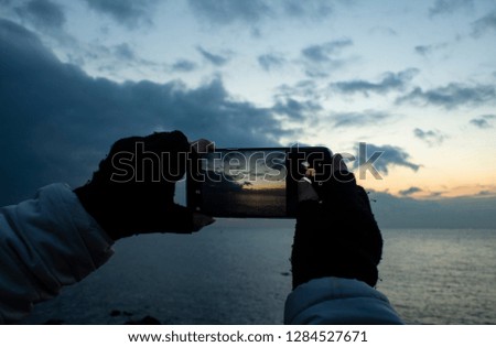 Girl's hand holding smartphone taking sunset photo. Hands in knitted gloves.