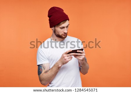 A man in a red cap looks into the phone in his hands and a tattoo on an orange background                   