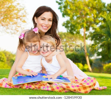 Picture of young happy family, beautiful mother with two cute kids having fun outdoors in spring, pretty female with son and daughter sitting down on green meadow on backyard, daycare concept