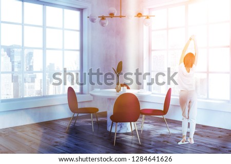 Woman in corner of modern dining room with white walls, wooden floor, round white table with red and gold chairs standing around it and two large windows. Toned image