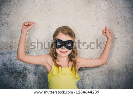 blond girl with black superhero mask and yellow shirt is posing in front of concrete background