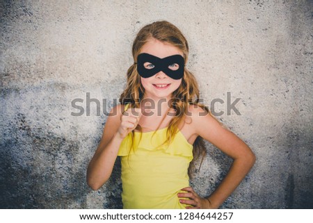 blond girl with black superhero mask and yellow shirt is posing in front of concrete background
