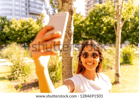A beautiful young woman in a white shirt and sunglasses standing next to a tree and taking a selfie.