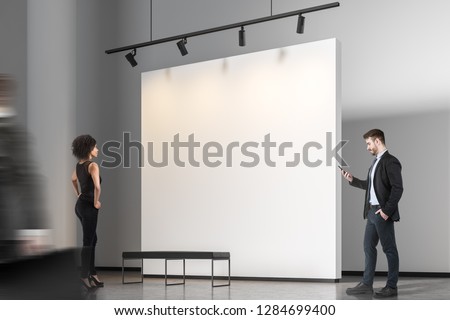 African American woman and bearded businessman with smartphone looking at blank gallery wall. Mock up blurred