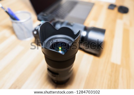 A DSLR lens on a wooden table together with a photo camera, batteries and a penholder.