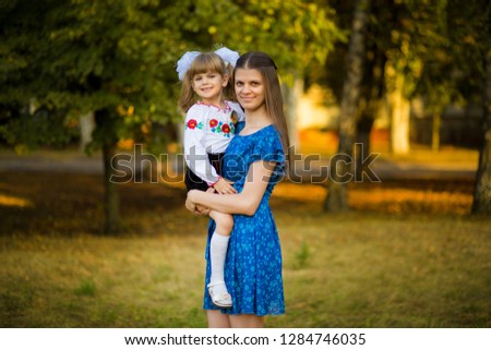 Portrait of beautiful young mother hugging first-grader daughter in festive school uniform on background autumn park.