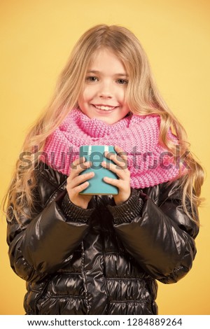 Tea or coffee break. Child hold mug in black jacket and pink scarf. Hot drink in cold weather. Autumn season relax concept. Girl with blue cup smile on orange background.