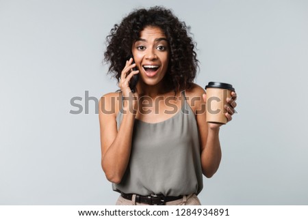 Happy young african woman casually dressed standing isolated over gray background, talking on mobile phone, holding takeaway cup of coffee