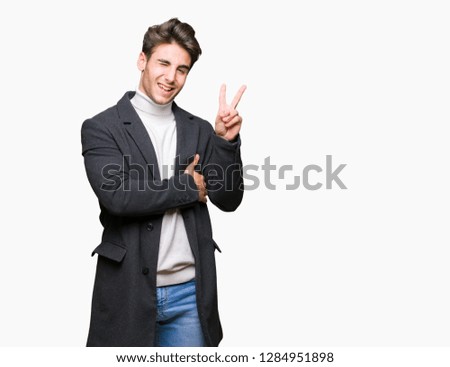 Young elegant man wearing winter coat over isolated background smiling with happy face winking at the camera doing victory sign. Number two.