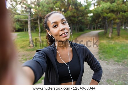 Portrait of pretty woman 20s wearing black tracksuit and earphones taking selfie photo on cell phone while walking through green park