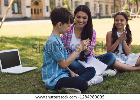 Teacher And Two Sudents Sitting On Grass In Park. Teacher Giving Apple To Boy.