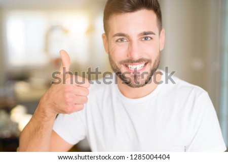 Young handsome man wearing casual white t-shirt at home doing happy thumbs up gesture with hand. Approving expression looking at the camera with showing success.