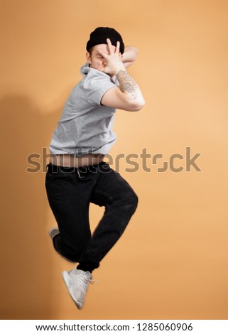 Stylish guy dressed in a gray shirt, black jeans and black hat with tattoo on his hand is jumping on the beige background in the studio
