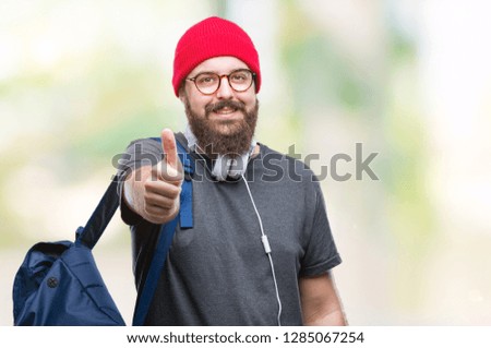 Young hipster man wearing red wool cap and backpack over isolated background doing happy thumbs up gesture with hand. Approving expression looking at the camera with showing success.