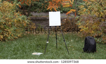 Easel, black backpack and paints are standing on the grass in the autumn park. Autumn concept