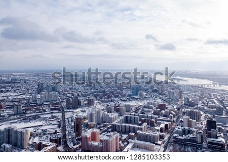 Winter landscape from a aerial view of the city of Novosibirsk in the haze, of the streets with a road, tall buildings, houses with roofs under white snow, a river, parking lots, cars, trees.
