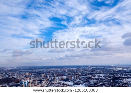 Scenic picture of the city in winter of a aerial, clear blue sky with white clouds, high-rise houses with roofs under the snow, streets with trees, industrial plants in the distance.