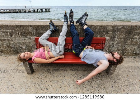 Young tired people friends with roller skates. Woman and man relaxing lying on bench outdoor.