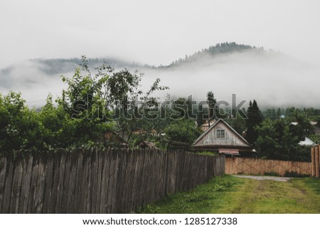 summer landscape panorama view of the village in the mountains early in the morning. to the right is a long wooden fence, fog in high mountains. green wood, on the sky white and gray clouds.