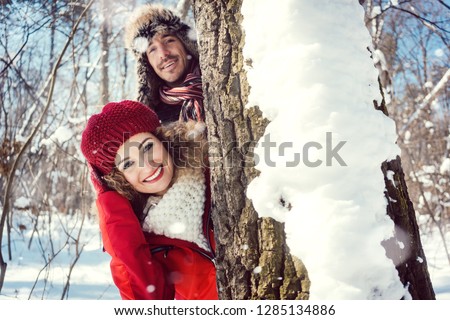 Playful couple hiding behind a tree trunk in the snow looking into the camera