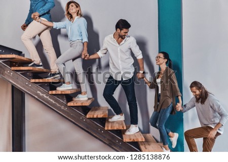 Achieving success together. Side view of young modern people in smart casual wear holding hands and smiling while moving up the stairs  Royalty-Free Stock Photo #1285138876