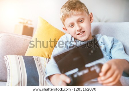 Preteen boy plays with smartphone connected with gamepad lying on the cozy sofa in the home living room