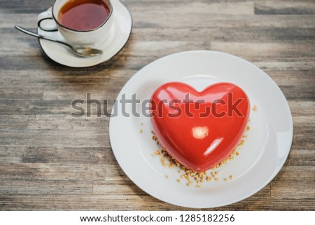 Red cake iced in the shape of a heart. Brown wooden table and cup of tea on the background. Valentine's Day holiday.