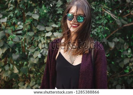 Portrait of happy boho girl in sunglasses smiling in sunny garden. Stylish hipster carefree girl posing on background of green bush in street. Space for text. Summer vacation