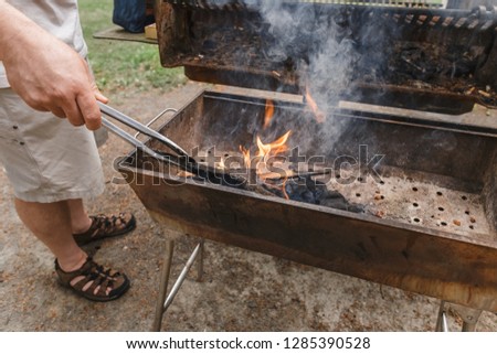 Man's hand preparing preparing for grilling in public outdoor charcoal grill in the summer park with wood ashes and fire flames