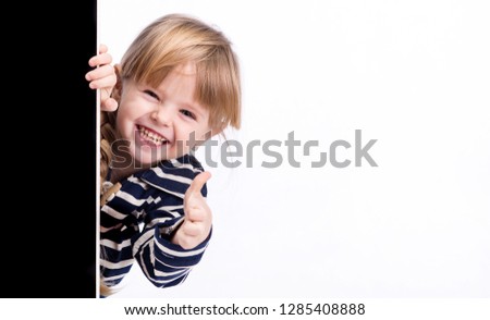 A little girl looks out from behind a black board. Shows finger gesture "good".
