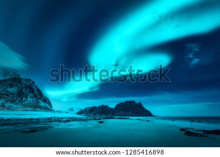 Beautiful aurora borealis. Northern lights in Lofoten islands, Norway. Starry sky with polar lights. Winter landscape with bright aurora, sea, sandy beach and snowy mountains. Uttakleiv beach at night