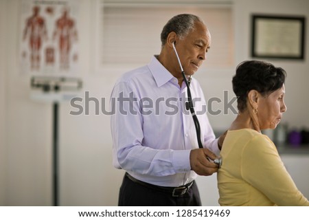 Male doctor listening to the heartbeat of a mature female patient inside his office. Royalty-Free Stock Photo #1285419469