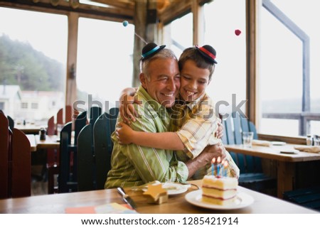 Smiling young boy and his father hugging next to a birthday cake while wearing funny hats inside a diner.