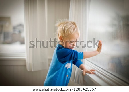 Young toddler pressing his hand against a window pane while standing inside a room.