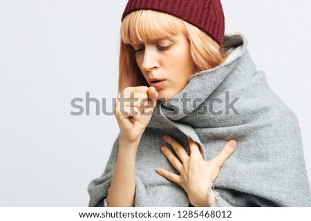 Cute sick young teen woman in red hat, wrapped in warm scarf coughing, closed eyes.Female feeling the first symptoms of illness.Bronchitis, upper respiratory infection concept.Moist cough, laryngitis Royalty-Free Stock Photo #1285468012
