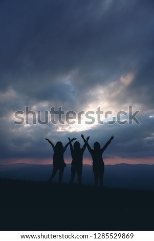 Silhouette image of three women standing and raising hands, watching sunset with mountains view in the evening