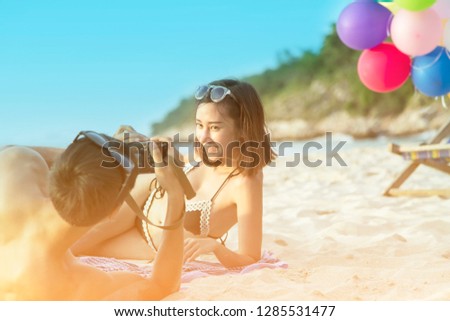 Happy Romantic Young Asian Couple Enjoying Beautiful Sunset Lying on the Beach. Cheerful Man Take A Photo For His Lovely Girl With Sunset Time. Travel Vacation, Valentine, Love, Lifestyle Concept.