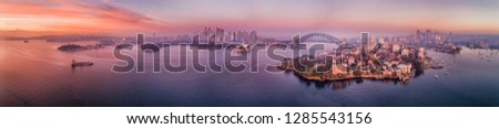 Red dawn sun light reflecting in waters of Sydney harbour and bays around waterfront of Sydney city CBD with landmarks in wide aerial panorama.