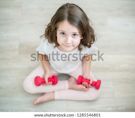 Little girl sitting on floor and lifting dumbbells, home workout. Kid doing exercise. Practicing yoga exercises. Healthy lifestyle, physical activity and fitness. Indoors. Looking at camera. Top view