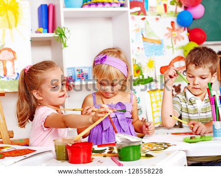 Child painting at easel in school. Teacher help. Royalty-Free Stock Photo #128558228
