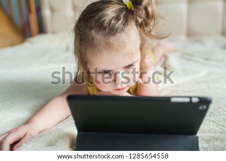 cute little happy girl is lying on the bed using looking digital tablet pad in the bedroom at home. family activity concept.