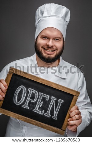 Handsome bearded man in white chef uniform smiling and showing chalkboard with open writing while standing on gray background