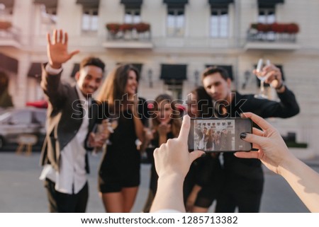 African man in long white shirt waving hand and smiling while his european female friend taking picture. Outdoor portrait of pleased people enjoying travel and posing to woman with black manicure.
