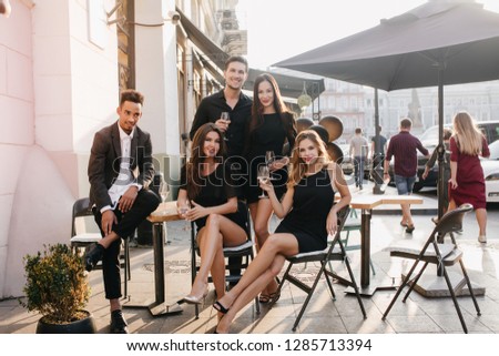 Stylish african man sitting in confident pose and looking at girls in outdoor cafe. Portrait of happy couple posing on the street with friends enjoying meeting.