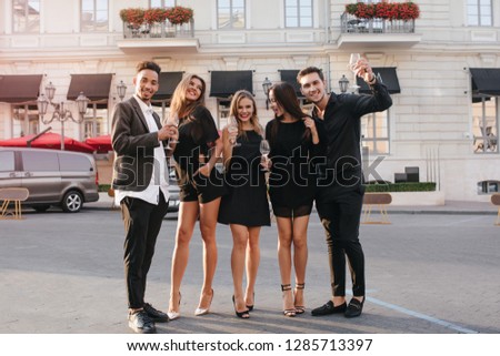 Pleased african man in white shirt and stylish shoes telling toast, enjoying wine with friends. Outdoor portrait of guys in formal suits and girls wears black dresses, posing on city background.