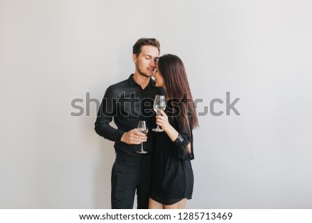 Long-haired brunette woman embracing her boyfriend while dancing at birthday party. Indoor photo of cute couple in black attires enjoying champagne and hugging on white background.