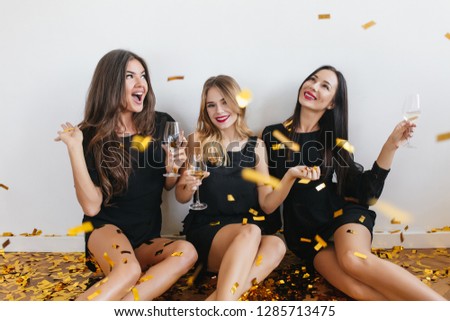 Brunette woman in black dress dreamy looking at confetti and gently smiling. Indoor portrait of lovely european girls relaxing at hen-party with wine and jokes.