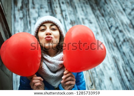 horizontal image of a cheerful young woman with a heart shaped red balloons. woman blows kiss, demonstrates her good feelings, says Love you on distance. Young pretty female model makes air kiss