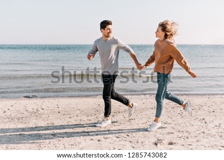 Blissful couple running down the beach in cold day. Outdoor photo of blithesome blonde girl fooling around at sea.