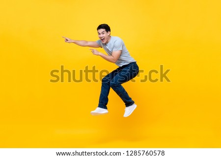 Energetic excited young Asian man jumping and pointing hand to empty space aside studio shot isolated on colorful yellow background